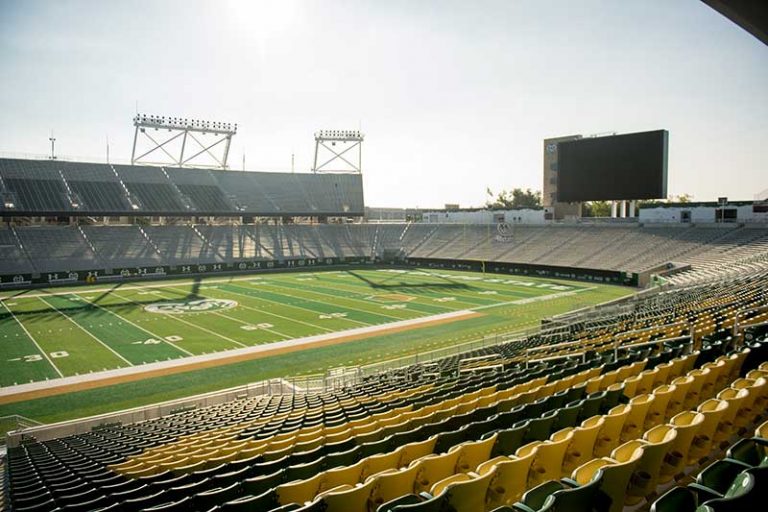 Canvas Stadium at Colorado State University, featuring green and gold seating, a large video scoreboard, and a bright, open view of the football field under clear skies.