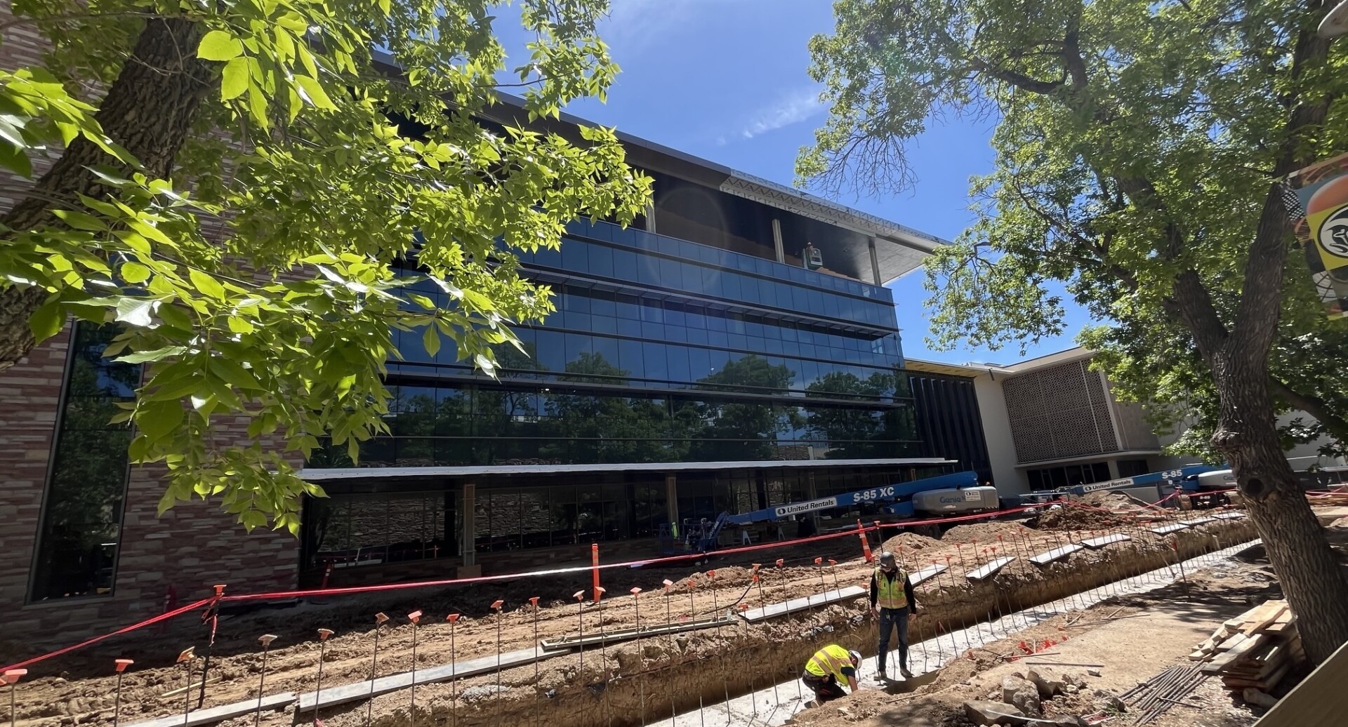 Construction activity outside the new Clark Building at Colorado State University, showing the glass exterior, active site work, concrete installation, and surrounding campus landscape improvements.
