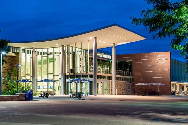 The Lory Student Center at Colorado State University, illuminated at dusk, featuring modern glass architecture, tall columns, and outdoor seating with umbrellas.